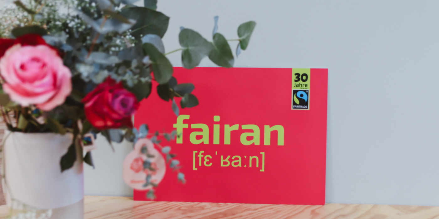 Rote Karte mit dem Wort "fairan" und einem Label "30 Jahre Fairtrade" auf einem Holztisch, daneben ein Blumenstrauß in einer Vase