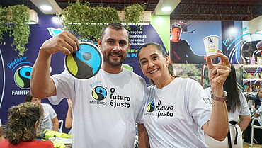 Ein junger Mann und eine junge Frau tragen weiße T-Shirts mit Fairtrade-Slogan und halten Fairtrade-Siegel in die Kamera. Im Hintergrund ist ein Messestand mit Fairtrade-Branding zu sehen.